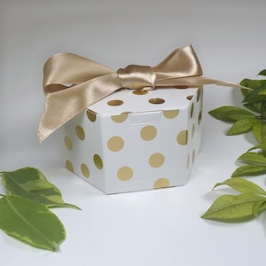 Könnte beinhalten: Eine weiße sechseckige Geschenkbox mit goldenen Punkten und einem goldenen Satinband, das zu einer Schleife gebunden ist.