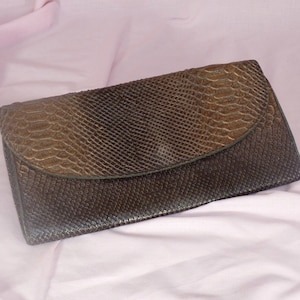 Peut inclure: Une pochette en cuir de serpent brun avec une fermeture à rabat. La pochette a une surface texturée et un léger brillant.