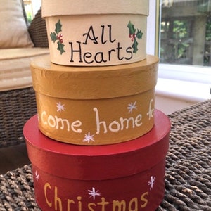 May include: Three round, painted, nesting boxes with lids. The top box is white with green holly and red berries and the text "All Hearts". The middle box is gold with white snowflakes and the text "come home for". The bottom box is red with white snowflakes and the text "Christmas".