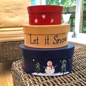 May include: Three round, painted wooden boxes stacked on top of each other. The top box is red with white stars. The middle box is tan with the words "Let it Snow" in black. The bottom box is blue with a snowman and two pine trees.