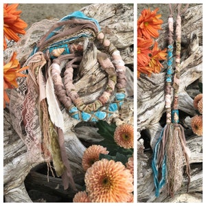 May include: A turquoise, brown, and white braided hair wrap with a tassel. The wrap is made of fabric and is tied around a piece of driftwood.