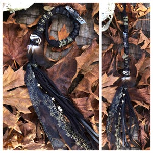 May include: Black and gold hair wrap with a crystal, feather, and beads. The wrap is on a bed of brown leaves.