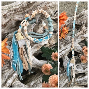May include: A blue and brown hair wrap with a white crystal charm and gold accents. The wrap is made of leather, suede, and fabric. It is tied around a piece of driftwood.
