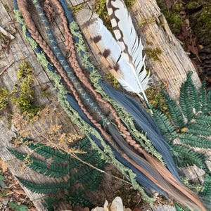 May include: A set of four braided hair extensions with brown, blue, and green strands. The extensions are tied together with a brown leather cord and have a frayed, rustic look. The extensions are displayed on a wooden surface with a feather and green foliage.