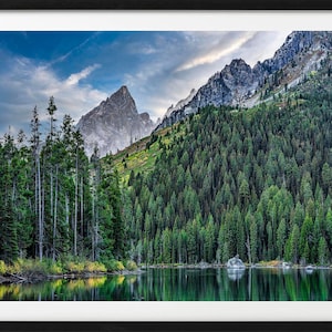 May include: A scenic landscape photograph featuring a mountain range with a lake in the foreground. The mountains are covered in lush green trees and the lake is reflecting the blue sky and clouds.