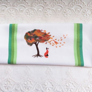 May include: A white kitchen towel with green stripes and an embroidered design of an orange fox looking up at a tree with orange and brown leaves blowing in the wind.