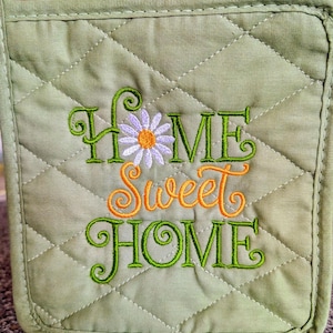 May include: A quilted, light green pot holder with the embroidered phrase "HOME SWEET HOME" in green and orange thread. A white daisy with an orange center is embroidered above the word "SWEET". The pot holder has a diamond pattern.