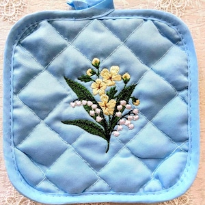 May include: A light blue quilted pot holder with a floral embroidery design. The embroidery features yellow flowers, white buds, and green leaves. The pot holder has a square shape with a loop for hanging.