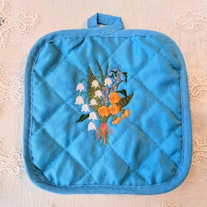 May include: A square, quilted, turquoise pot holder with a loop for hanging. The center features an embroidered bouquet of white lily of the valley, orange and blue flowers, and green leaves.