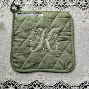 May include: A square, olive green quilted pot holder with a cream-colored embroidered letter "K" and floral design. The pot holder has a small loop for hanging and a stitched border. The website address "KateKaminskiBlog.com" is printed on a small tag.