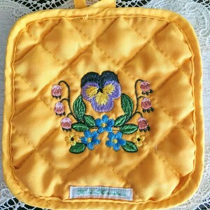 May include: Yellow quilted pot holder with embroidered floral design. The embroidery features a purple and yellow pansy, blue flowers, and pink bell-shaped flowers. A small label with text is at the bottom. The pot holder has a hanging loop.