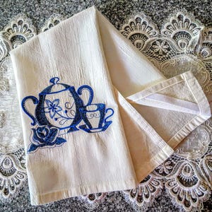 May include: Cream-colored tea towel with a blue embroidered design of a teapot, teacup, and rose. The towel is folded, revealing the embroidered design. The background is a textured surface with lace trim.