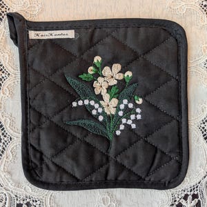 May include: Black quilted pot holder with a floral embroidery design. The embroidery features white flowers, green leaves, and small white buds. The pot holder has a loop for hanging and a tag that reads "KaiaKanvas".