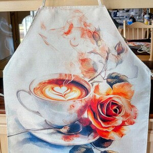 May include: A white apron with a watercolor design of a cup of coffee with a heart-shaped design in the foam and a red rose with green leaves.