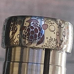 May include: A silver ring with an engraved design of two sea turtles and coral. The turtles are outlined in black and have a brown shell pattern. The coral is white and has a detailed design.