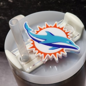 May include: White tumbler with a clear lid and a straw. The lid has a clear plastic attachment with a Miami Dolphins logo featuring a blue dolphin and an orange sun.