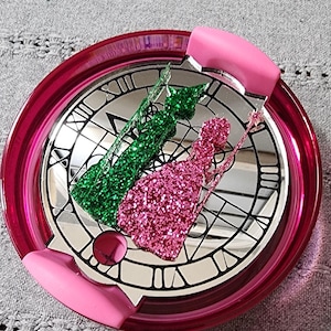 May include: A pink and green glitter silhouette of two figures, a princess and a fairy, on a mirrored clock face with a pink lid.