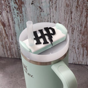 May include: A light green travel mug with a black and white Harry Potter logo on the lid. The mug has a stainless steel band and a black handle.