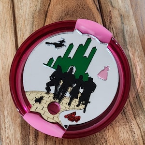 May include: A red and pink round lid with a white background featuring a green Emerald City, a yellow brick road, a black dog, and a pink witch. The characters from the Wizard of Oz are walking on the yellow brick road.