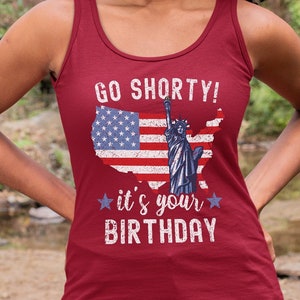 Puede incluir: Camiseta de tirantes roja con un dise&ntilde;o de bandera estadounidense desgastado y el texto "Go Shorty! It's your Birthday" en blanco.