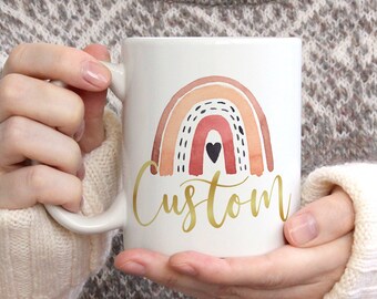 May include: White ceramic mug with a watercolor rainbow design in shades of pink, orange, and brown. The rainbow has a small black heart in the center. The word "Custom" is written in gold script below the rainbow.