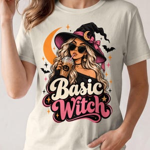 May include: A cream-coloured t-shirt featuring a graphic of a blonde character wearing a black witch hat with a pink bow, sunglasses, and a choker. The character holds an iced coffee with a pumpkin design. The text Basic Witch is printed below.