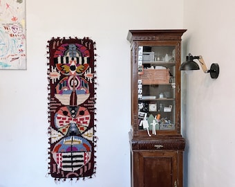 Puede incluir: Un armario de madera oscura con puertas de vidrio y estantes llenos de diversos objetos. Un tapiz de pared con un diseño geométrico colorido cuelga en la pared a la izquierda del armario. Una lámpara de pared con una pantalla negra se encuentra a la derecha del armario.