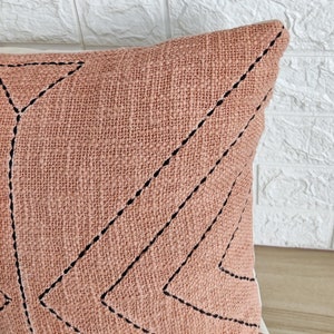 May include: A blush pink throw pillow with a geometric pattern of black stitched lines forming triangles. The pillow is made of a textured fabric.