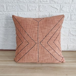 May include: A peach-colored throw pillow with a geometric pattern of black stitching. The pillow is made of a textured fabric and has a square shape.