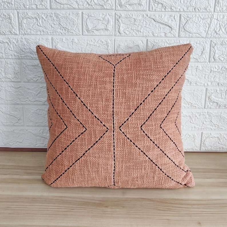 May include: A peach-colored throw pillow with a geometric pattern of black stitching. The pillow is made of a textured fabric and has a square shape.