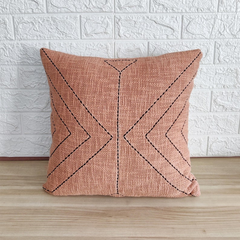 May include: A peach-colored throw pillow with a geometric pattern of black stitching. The pillow is made of a textured fabric and has a square shape.