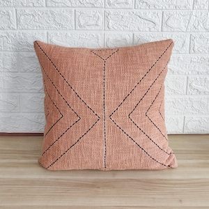 May include: A peach-colored throw pillow with a geometric pattern of black stitching. The pillow is made of a textured fabric and has a square shape.