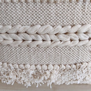 May include: A close-up of a cream-colored woven wall hanging with a fringe border. The hanging has a textured pattern and is made of natural fibers.