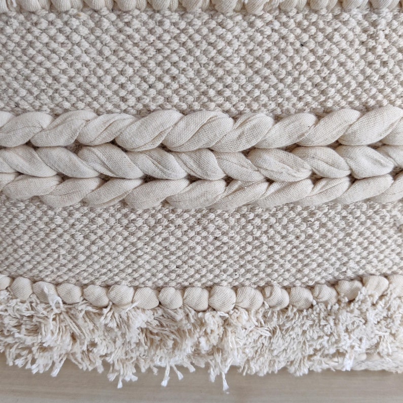 May include: A close-up of a cream-colored woven wall hanging with a fringe border. The hanging has a textured pattern and is made of natural fibers.
