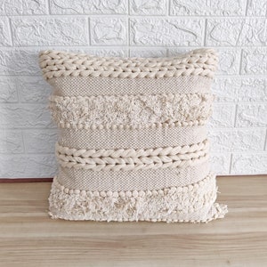 May include: A cream-colored throw pillow with a woven texture and fringe detailing. The pillow has a braided border and a series of horizontal bands with pom-poms.