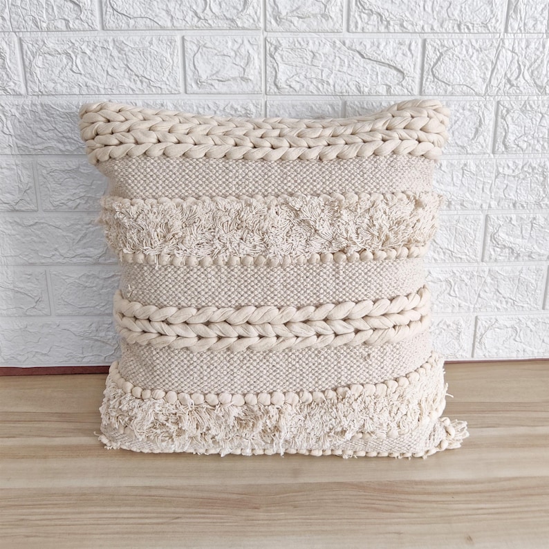 May include: A cream-colored throw pillow with a woven texture and fringe detailing. The pillow has a braided border and a series of horizontal bands with pom-poms.