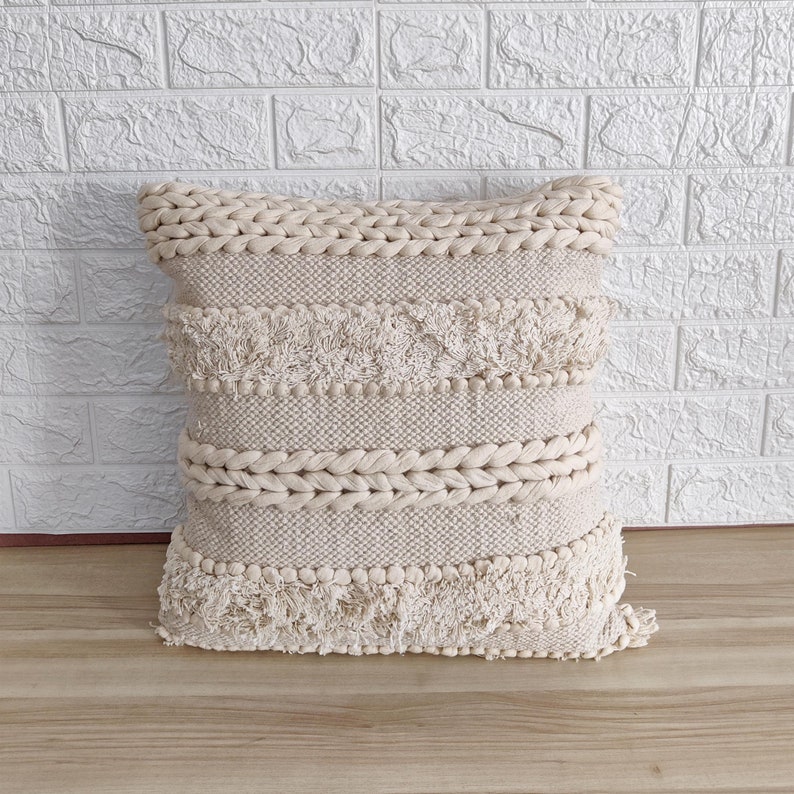May include: A cream-colored woven throw pillow with a fringe border and a braided design. The pillow is made of a soft, textured fabric and has a subtle, natural look.