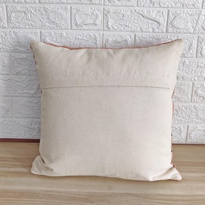May include: A cream-colored throw pillow with a zipper closure on the back. The pillow has a peach-colored border and is made of a soft, woven fabric.