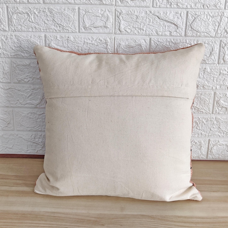 May include: A cream-colored throw pillow with a zipper closure on the back. The pillow has a peach-colored border and is made of a soft, woven fabric.