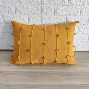 May include: A mustard yellow throw pillow with a textured fabric and decorative pom-poms around the edges.