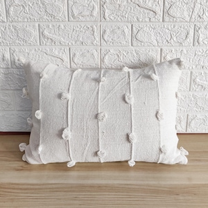 以下が含まれることがあります： 縁に小さな白いポンポンが付いた、白の矩形のクッション。テクスチャのある生地を使用しています。