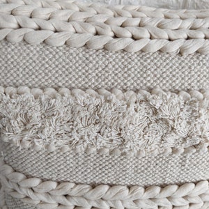 May include: A cream-colored throw pillow with a woven texture and a fringe border. The pillow has a unique design with different textures and patterns.