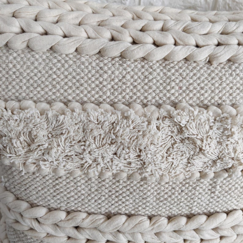 May include: A cream-colored throw pillow with a woven texture and a fringe border. The pillow has a unique design with different textures and patterns.