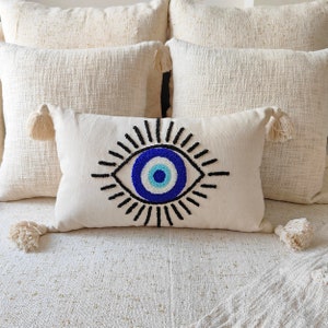 May include: A white throw pillow with a blue and white evil eye design. The evil eye is embroidered with black thread and has a blue, white, and light blue center.