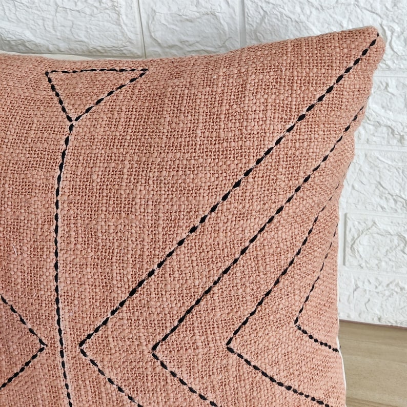 May include: A blush pink throw pillow with black geometric stitching. The pillow has a textured fabric and a subtle sheen.