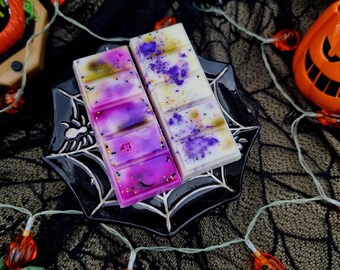 May include: Two wax melts in a black spiderweb design dish. The wax melts are white with purple and gold glitter. The dish is on a black lace background with orange pumpkin lights.