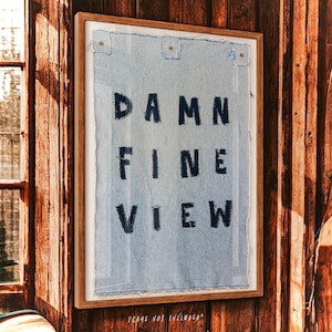 Puede incluir: Obra de arte enmarcada con las palabras "DAMN FINE VIEW" en letras negras sobre una tela azul claro. La obra está montada en un marco de madera y está fijada a una pared de madera. El texto "FRAME NOT INCLUDED" está en la parte inferior.