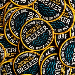 Könnte beinhalten: Ein Haufen schwarzer und goldfarbener gestickter Aufnäher. Jeder runde Aufnäher hat einen goldenen Rand und die Worte "BREAKER" und "TIDE WAITS FOR NO ONE". Die Aufnäher enthalten auch den Text "DO THE THING NOW AOTEAROA NEW ZEALAND".