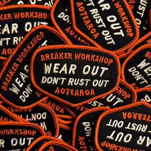 May include: A collection of black embroidered patches with orange borders. The patches feature the text "BREAKER WORKSHOP WEAR OUT DON'T RUST OUT AOTEAROA" in white and orange thread. The patches are oval-shaped.