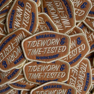 Könnte beinhalten: Ein Haufen brauner, gestickter Aufnäher mit dem Text "Tideworn & Time-Tested Workshop" in Weiß und Blau. Die Aufnäher haben einen cremefarbenen Rand, und das Wort "Breaker" ist ebenfalls sichtbar.